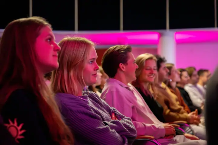 Rij jonge bezoekers in warm, roze licht luistert met een glimlach naar de spreker