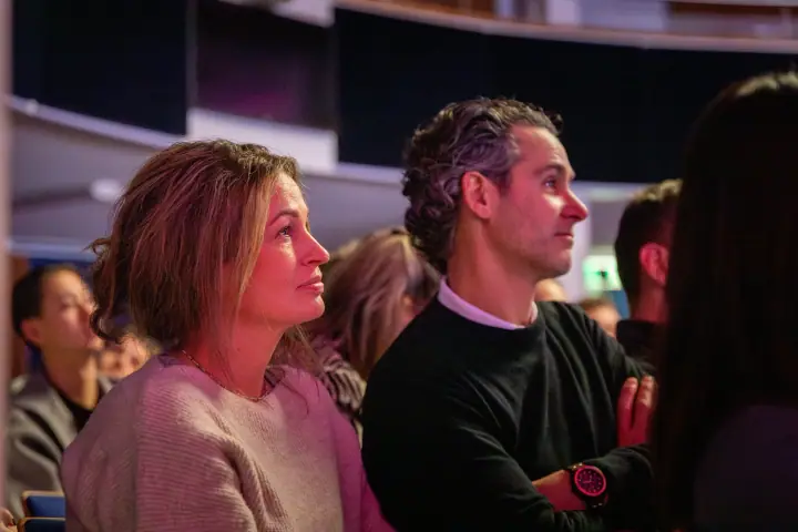Vrouw en man kijken geboeid omhoog naar het podium, hij met de armen over elkaar