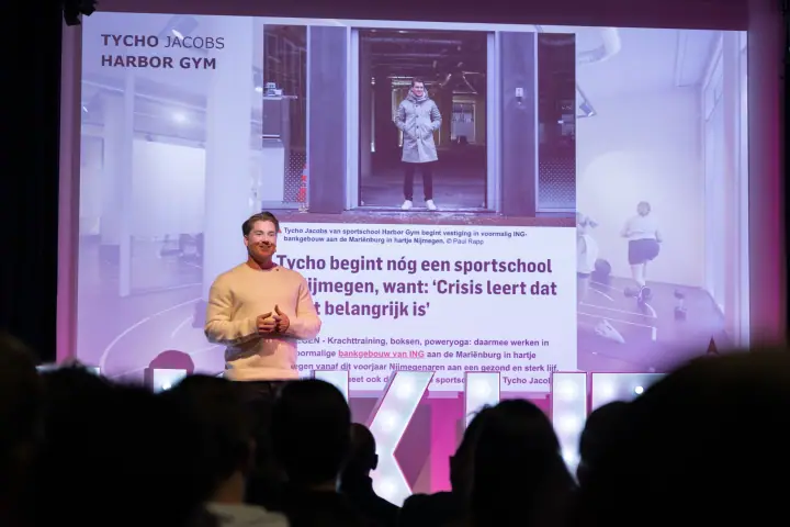 Lachende spreker in beige trui voor zijn slide, het publiek als donkere silhouetten op de voorgrond