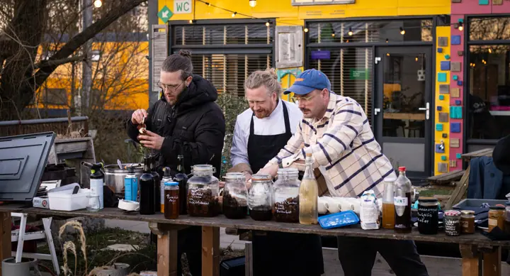 Drie mannen aan een buitentafel vol potten en flessen, voor een felgeel gebouw