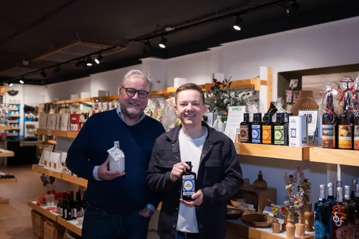Twee mannen in een winkel houden elk een fles Virnelli olijfolie vast en lachen naar de camera