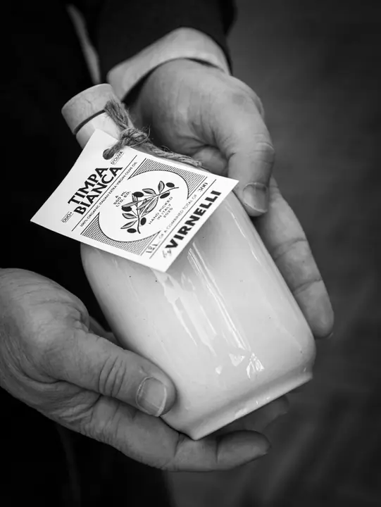 Zwart-wit: handen houden een stenen fles Timpa Bianca olijfolie met het label van Virnelli