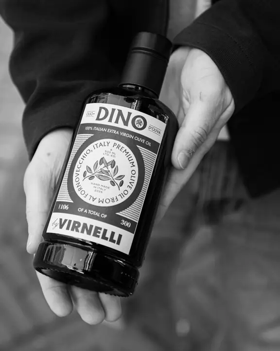 Zwart-wit productbeeld van een fles DINO olijfolie, voorzichtig vastgehouden in twee handen